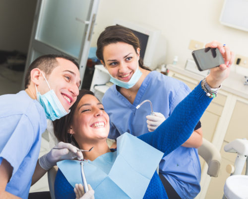 Happy Patient, Dentist and Assistant Taking Selfie All Together. Patient is Holding Smart Phone, Dentist and Assistant are Holding their tools. Focus on Patient Eyes